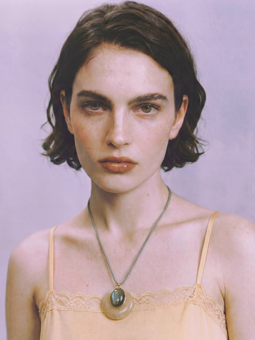 Model wearing the MOUNSER Aarnio Pendant in Olive Oil with yellow jade and labradorite disc pendant on twisted cord, styled with a blush lace camisole against a lavender background.