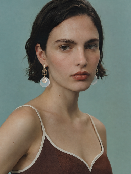 Model wearing MOUNSER Crespi Earrings in Moonlight with opalite disc and cubic zirconia teardrop charms, styled with a rust camisole against a teal background.