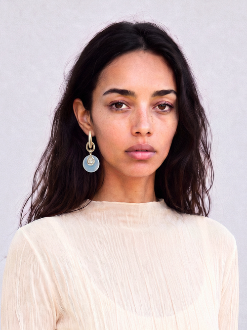 Model wearing MOUNSER Crespi Earrings in Moonlight with opalite disc and cubic zirconia teardrop charms, styled with a rust camisole against a teal background.