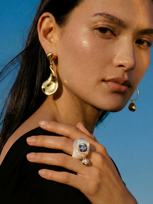 Model wearing MOUNSER Caspian Ring in Celeste Opal - white resin ring with opalite glass and gray Austrian crystal teardrop charm styled with sculptural gold MOUNSER Lunar earrings against blue sky