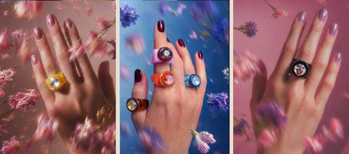 Three hands wearing MOUNSER Seamless Spectrum Rings in yellow, purple, orange, blue, tortoise and black resin colorways with iridescent Swarovski crystal gemstones, styled with dried florals in pink and blue, handcrafted art object jewelry made in Los Angeles.
