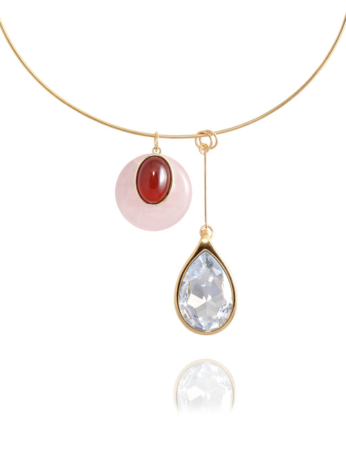 Close-up detail of the MOUNSER Rega Collar Necklace in Amarena showing the pink and red carnelian domed pendant above an elongated clear Austrian crystal teardrop in 14k gold-plated brass setting, with reflection.
