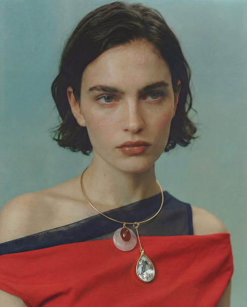 Model wearing the MOUNSER Rega Collar Necklace in Amarena with pink and red carnelian dome pendant and crystal teardrop, styled with a red draped top against a teal background.