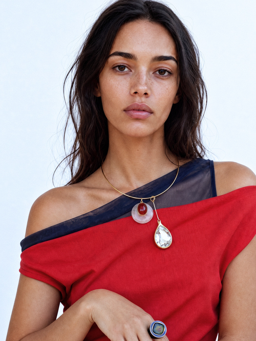 Close-up detail of the MOUNSER Rega Collar Necklace in Amarena showing the pink and red carnelian domed pendant above an elongated clear Austrian crystal teardrop in 14k gold-plated brass setting, with reflection.