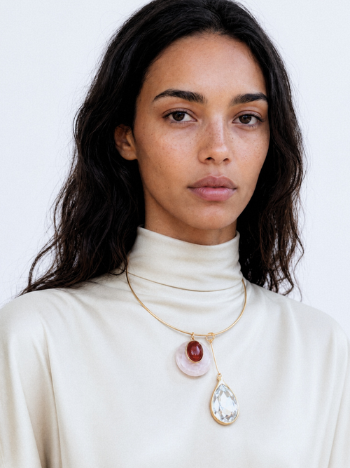 Model wearing the MOUNSER Rega Collar Necklace in Amarena with brass collar and pink quartz and red carnelian dome pendant and crystal teardrop, styled with a white draped top against a teal background.
