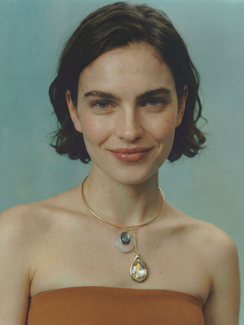 Model wearing the MOUNSER Rega Collar Necklace in Champagne with white quartz and crystal pendants, styled with a rust strapless top against a teal background.
