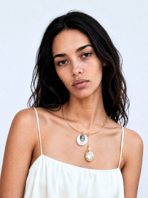 Model wearing the MOUNSER Rega Collar Necklace in Champagne with white quartz and crystal pendants, styled with a rust strapless top against a teal background.