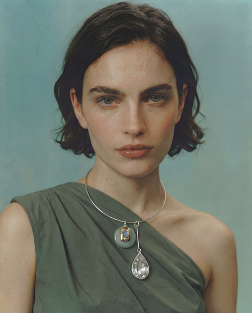 Model wearing the MOUNSER Rega Collar Necklace in Cucumber with jade and crystal pendants, styled with a sage green one-shoulder top against a teal background.