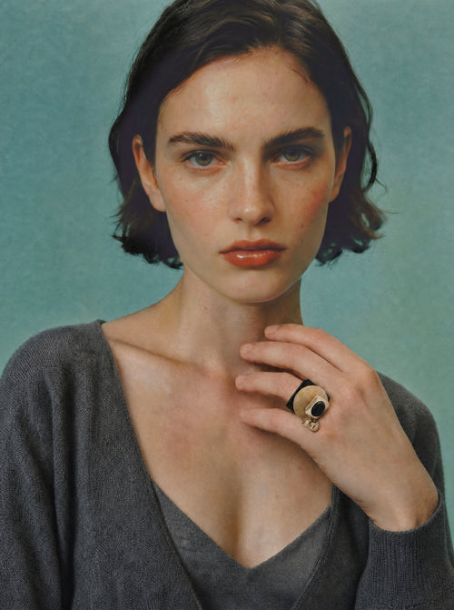 Model wearing the MOUNSER Rizzo Ring in Caramel styled with a grey v-neck top against a teal background.