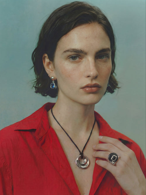 Model wearing the MOUNSER Rizzo Ring in Ink Lacquer alongside a coordinating necklace, styled with a red button-down shirt against a teal background.