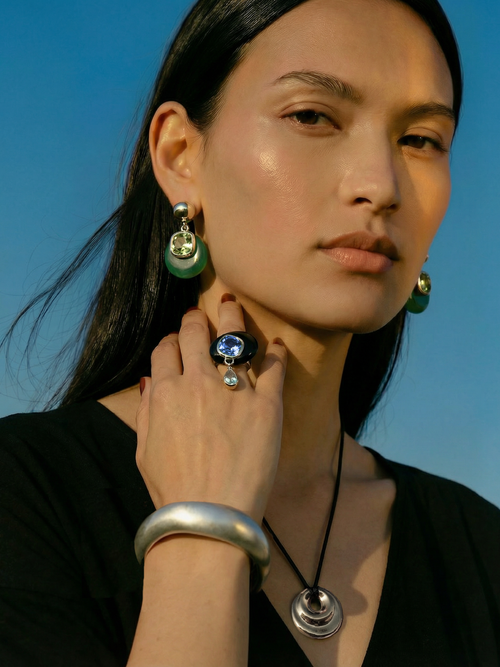 Model wearing MOUNSER Caspian Ring in Abyss - clear acrylic ring with black obsidian and sapphire crystal teardrop charm styled with Isle Earrings in Comber Green silver cuff and MOUNSER necklace against blue sky