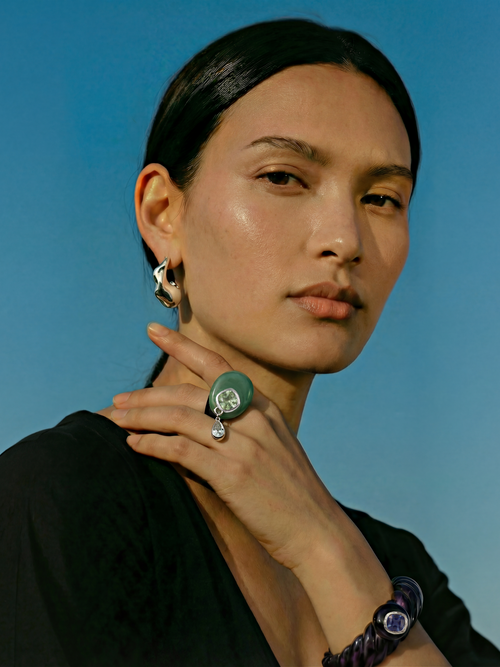 Model wearing MOUNSER Caspian Ring in Comber Green - black resin ring with green jade disc and crystal teardrop charm styled with sculptural silver MOUNSER earrings and purple bracelet against blue sky