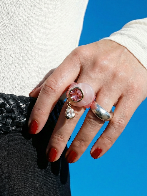Close up of MOUNSER Caspian Ring in Conch Pink - pink rose quartz resin ring with deep pink Austrian crystal and dangling cubic zirconia teardrop charm styled with sculptural silver ring red nails and white sweater against blue sky