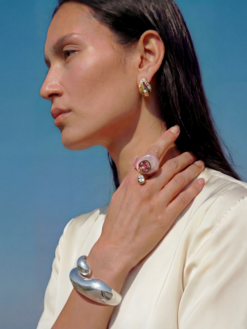 Model wearing MOUNSER Caspian Ring in Conch Pink - pink rose quartz resin ring with crystal teardrop charm styled with gold MOUNSER earrings and sculptural silver cuff against blue sky