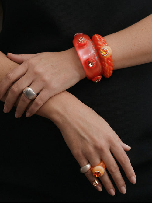 Model wearing MOUNSER Caspian Ring in Papaya - orange resin ring with citrus crystal and teardrop charm styled in warm tonal stack with Scallop Cuff bracelets and silver ring against black background