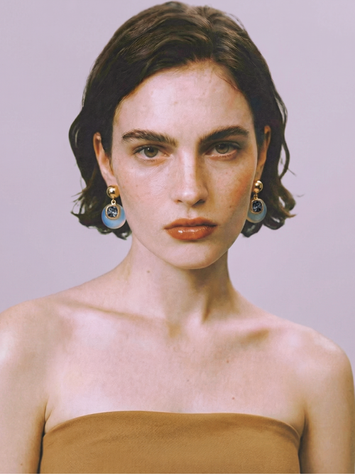 Model wearing MOUNSER Isle Earrings in Celeste Opal - opalite glass and gray Austrian crystal drop earrings with gold ball post styled with camel strapless top against lavender background