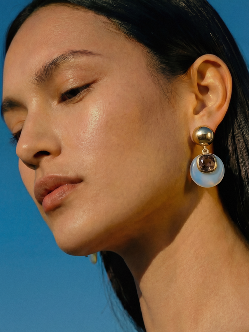 Close up profile of model wearing MOUNSER Isle Earrings in Celeste Opal - sculptural opalite glass disc and gray cushion-cut Austrian crystal drop earrings with 14k gold-plated ball post against blue sky