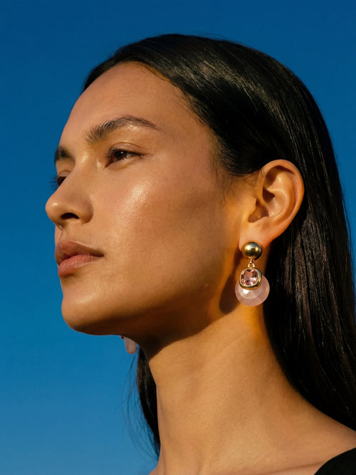 Model wearing MOUNSER Isle Earrings in Conch Pink - sculptural rose quartz and pink Austrian crystal drop earrings with gold ball post against blue sky
