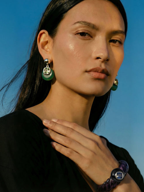 Model wearing MOUNSER Scallop Cuff in Abyss - deep purple resin cuff with sapphire crystal styled with MOUNSER Isle Earrings in Comber Green and hand at neck against blue sky