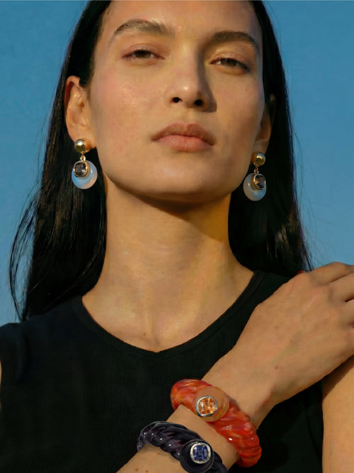 Model wearing MOUNSER Scallop Cuff in Papaya and Abyss bracelet stack - orange red and purple resin cuffs with crystals styled with Isle Earrings in Celeste Opal against blue sky