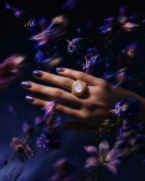 MOUNSER Spectrum Ring in Foundation worn on model's hand — milky beige limited edition cocktail ring with iridescent Austrian crystal on a dark purple floral background,  art object jewelry handcrafted to order in Los Angeles.